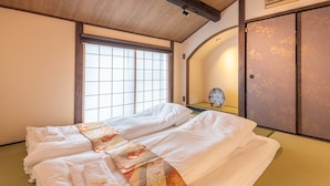 A traditional Japanese-style room with tatami flooring, a low bed with white bedding, a shoji screen door, and a small decorative table. - KumoMachiya Goshohigashi (Kyoto)