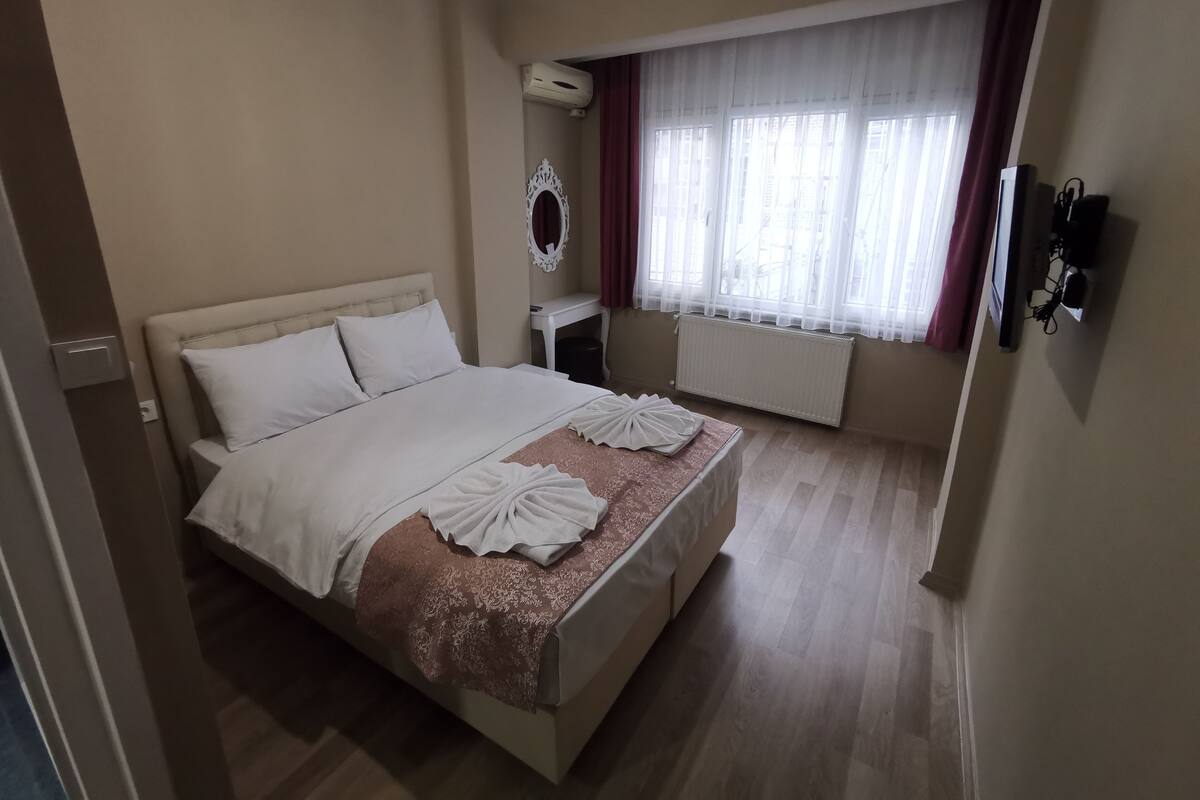 A hotel room with a bed, a bedside table, a mirror, a window with curtains, and a wall-mounted TV.