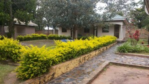A courtyard with a stone pathway, yellow flowering bushes, and a building with a tiled roof. - Blue Dove Hotel (Koboko)