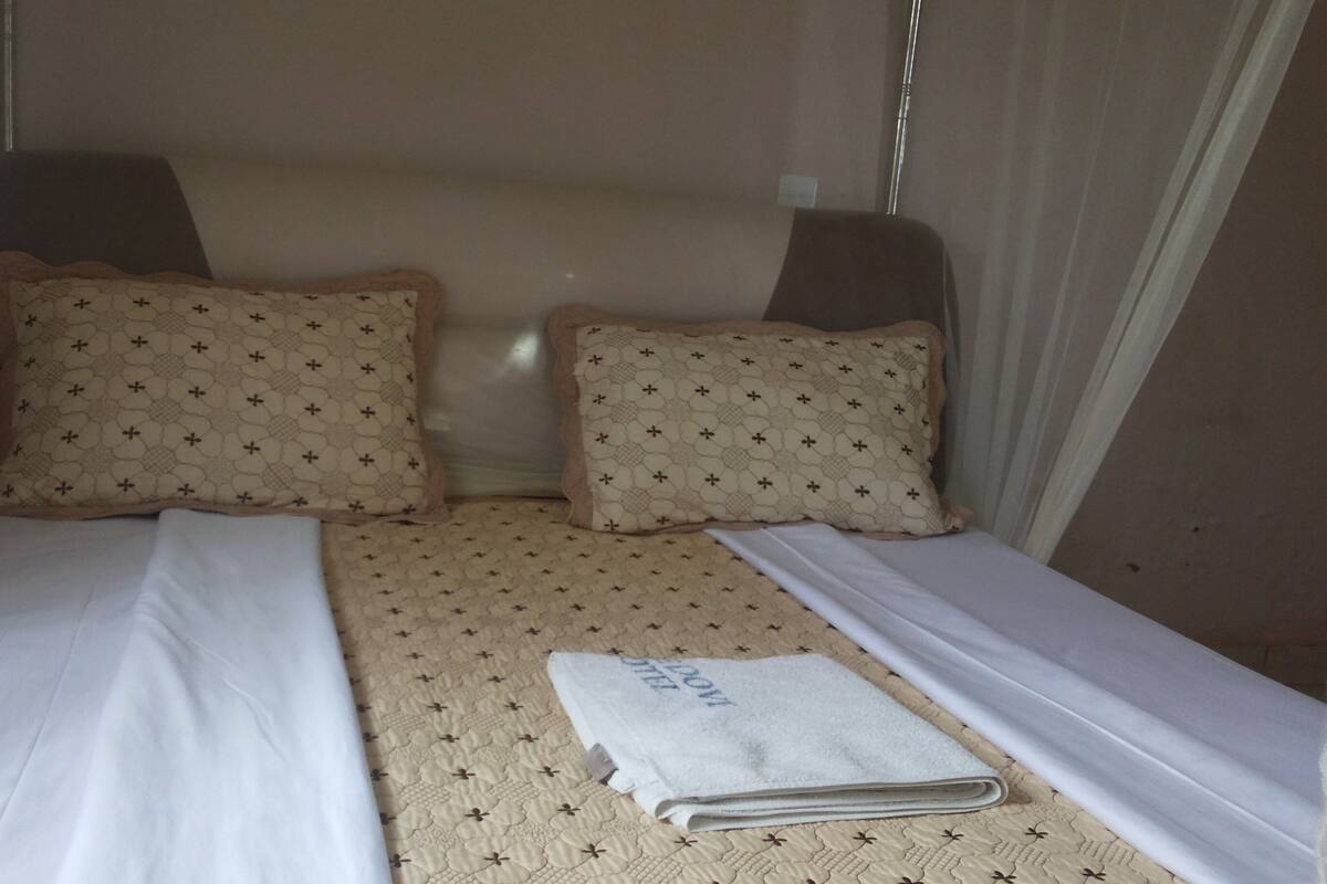 Una cama individual con una colcha estampada y dos almohadas, una toalla doblada y cortinas traslúcidas.
