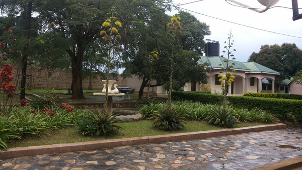A garden with a fountain, stone pathway, and a building with a green roof. - Blue Dove Hotel (Koboko)