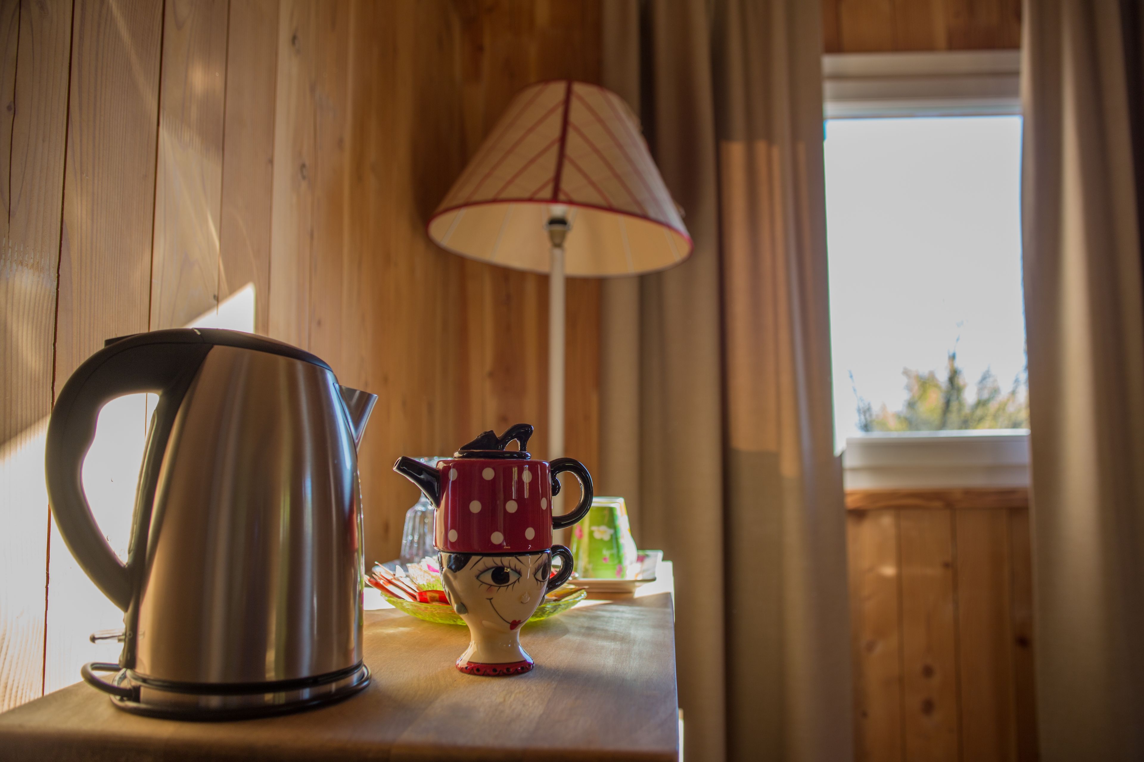 A wooden table with a stainless steel kettle, a polka-dotted mug, and a floor lamp.