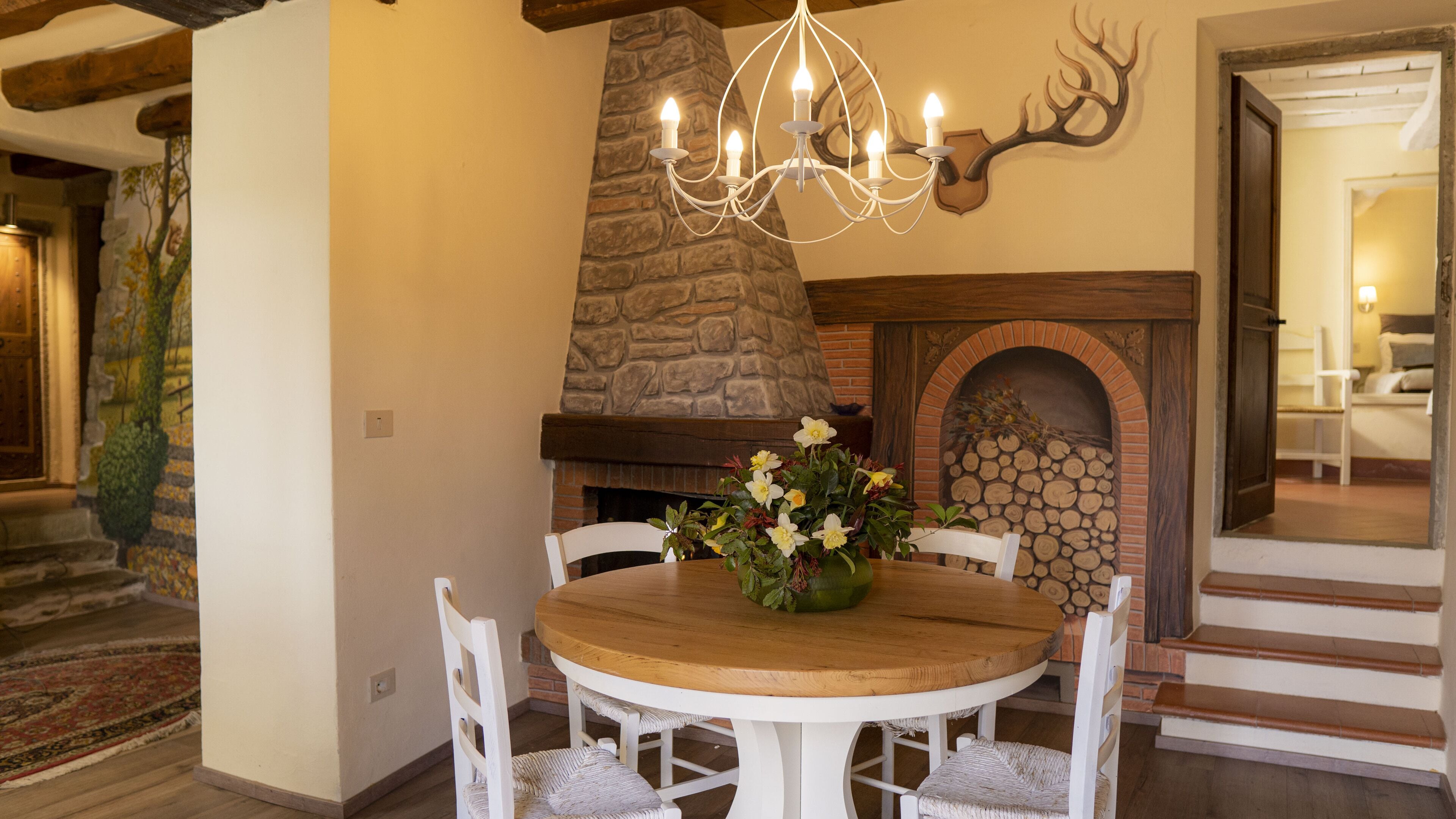 Ein Essbereich mit einem runden Holztisch, weißen Stühlen, einer Kristallleuchter, einem Steinkamin und einer Dekoration aus Hirschhörnern an der Wand.