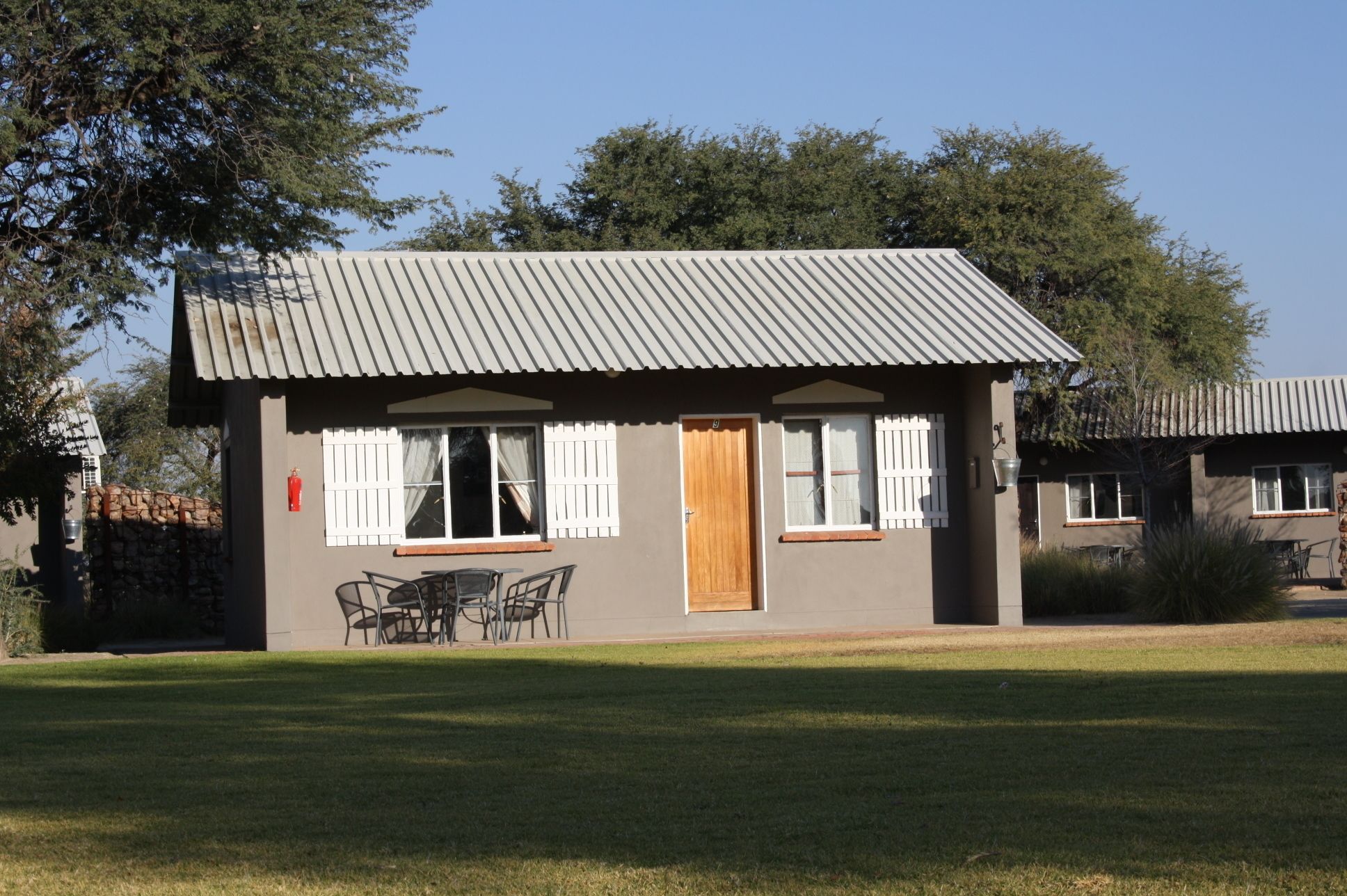 A small, single-story building with a metal roof, a wooden door, and a window with shutters. There is a table and chairs outside.