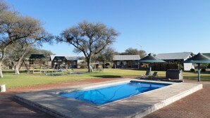 A swimming pool with a stone border, surrounded by a grassy area with trees and a pavilion. - OUT OF Nature Country Lodge (Windhoek)