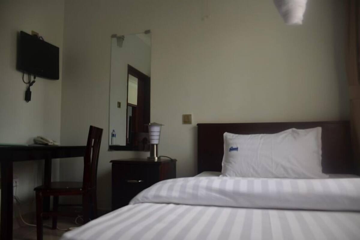 A hotel room with a bed, a desk, a chair, a bedside table, a lamp, and a TV mounted on the wall.