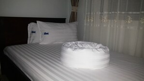 Ein ordentlich bezogenes Bett mit gestreifter Bettwäsche und zwei Kissen, ein runder, weißer Hocker und durchscheinende Vorhänge mit einem Blumenmuster.