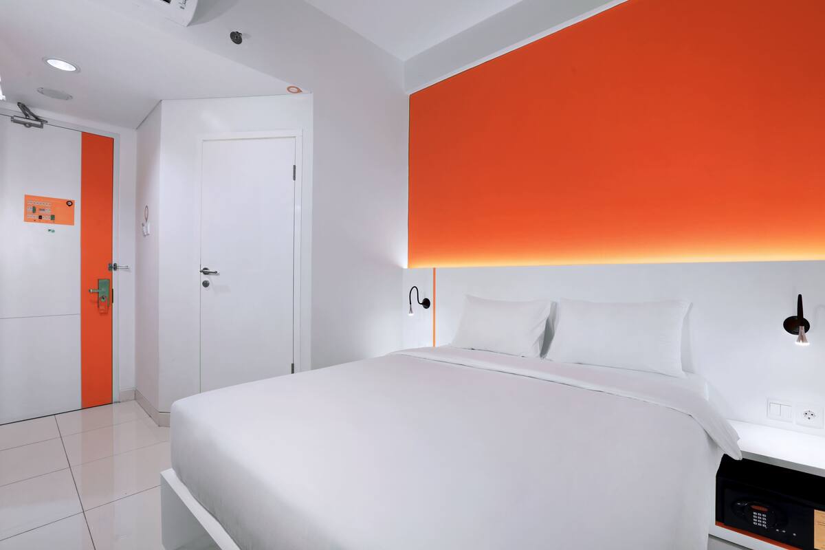 A modern hotel room with a large bed, a white door with an orange stripe, and a bright orange accent wall.