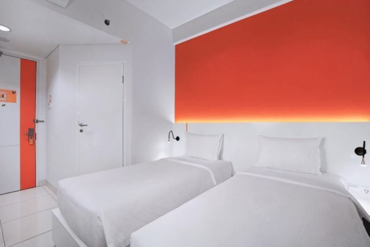 Une chambre d’hôtel avec deux lits, une porte blanche et un mur d’accent rouge.