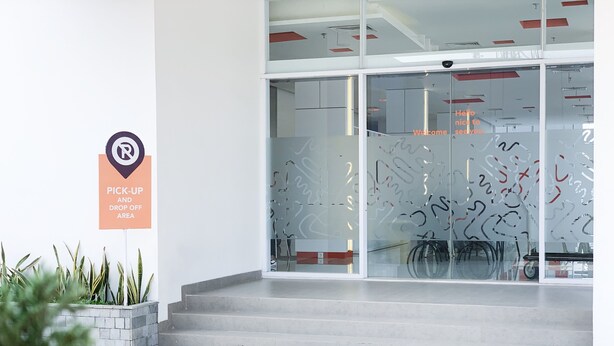 Entrada moderna de un edificio con puerta de vidrio, zona de recogida y descenso de viajeros, y una cámara de seguridad sobre la puerta.