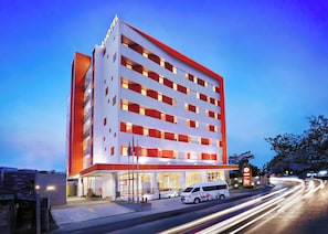 Un hotel de varios pisos con fachada moderna, destacando una entrada principal y una furgoneta estacionada frente a él.