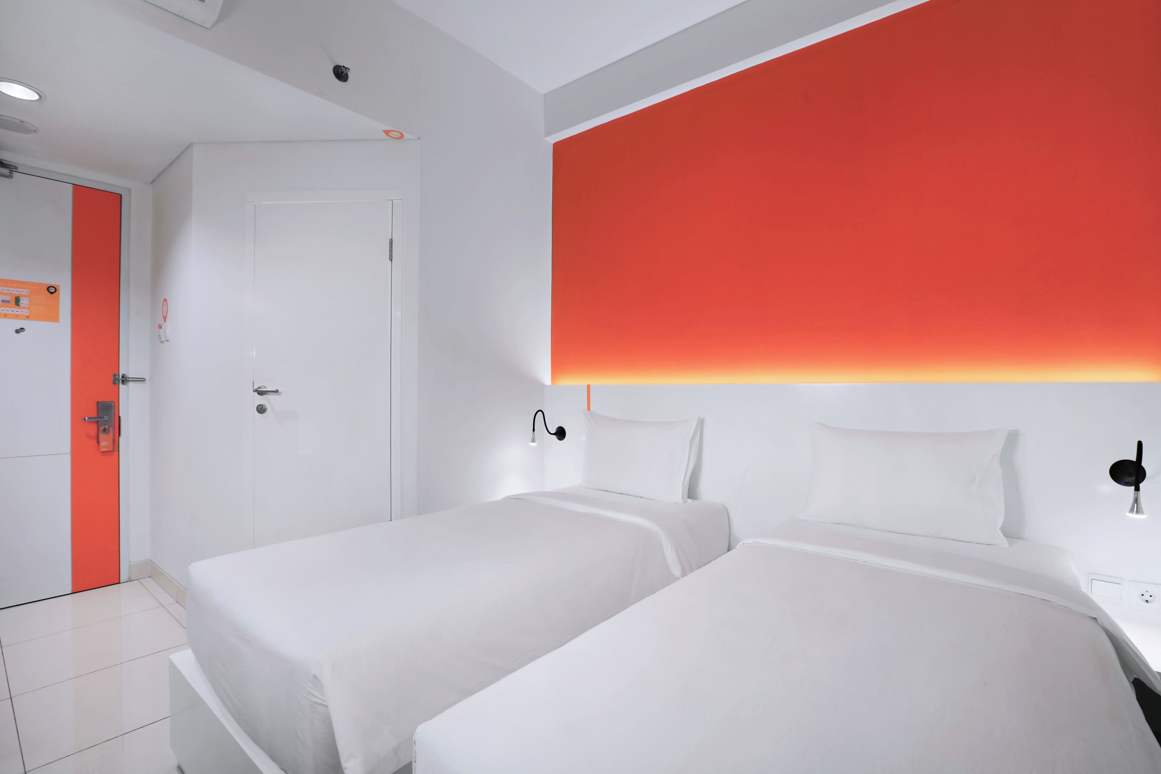Une chambre d’hôtel avec deux lits, une porte blanche et un mur d’accent rouge.