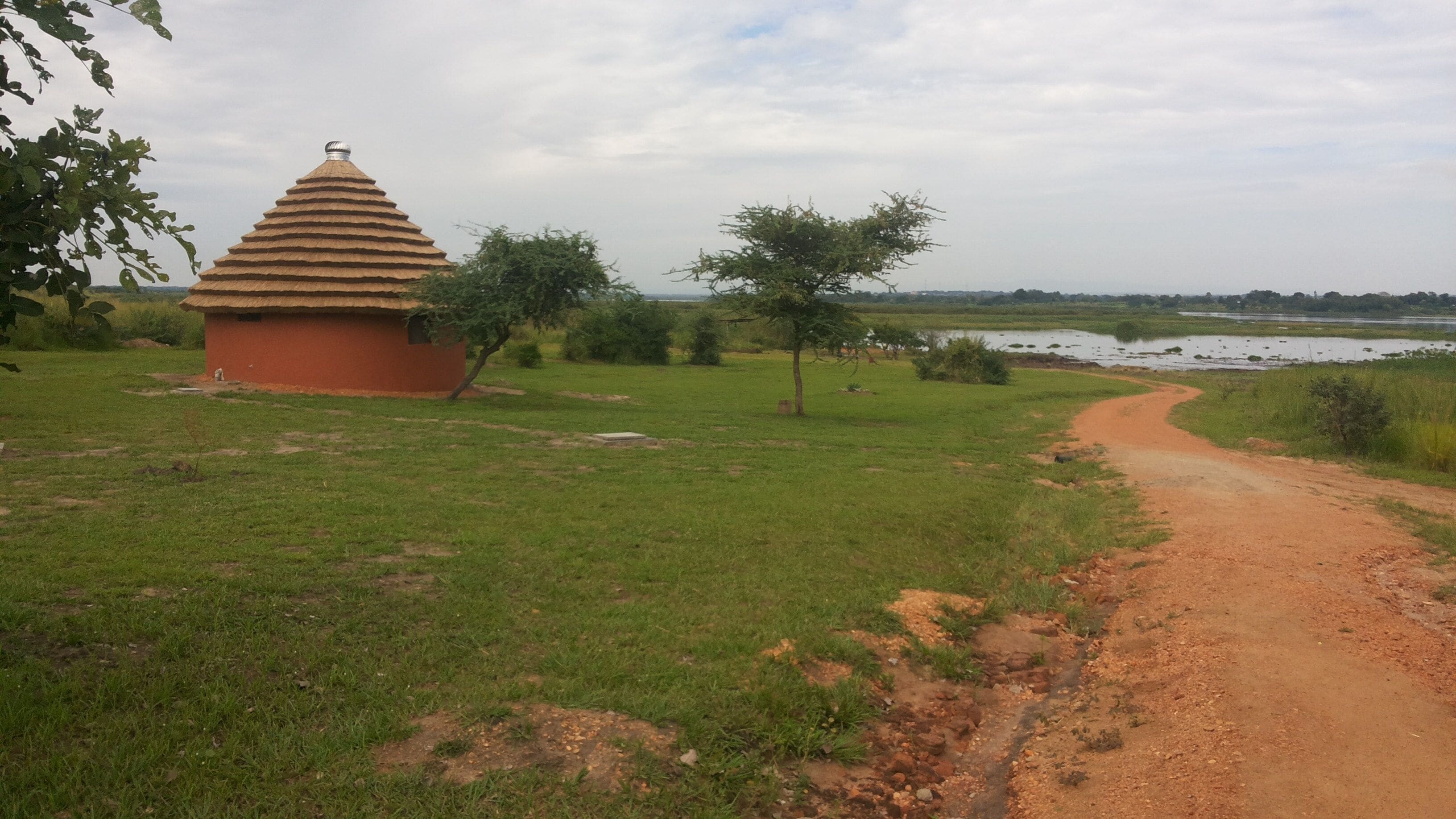 A rural landscape with a dirt path leading to a small red building with a conical roof, surrounded by greenery and a body of water in the background.
