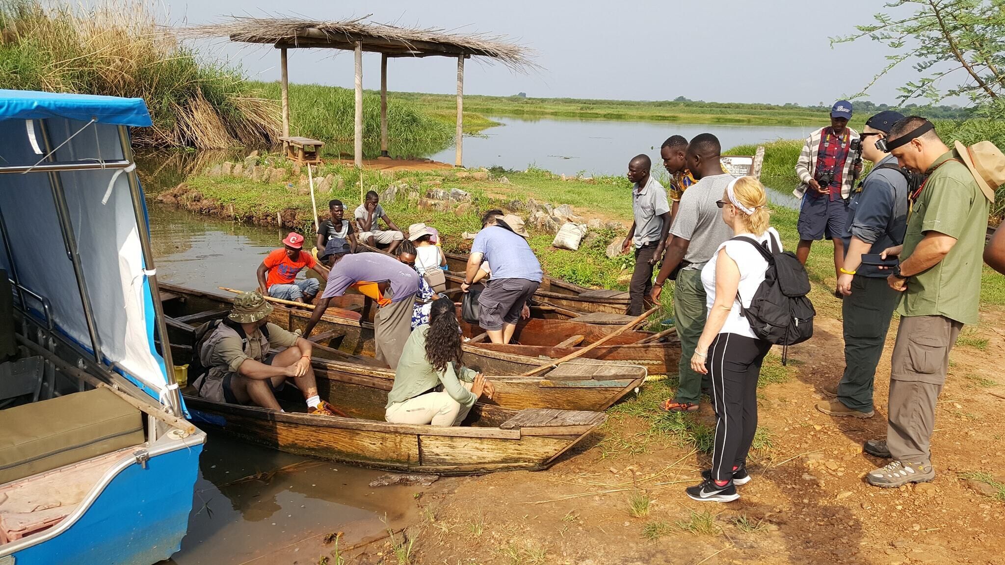 A group of people around a docked boat, some standing and others sitting, with a thatched shelter in the background.