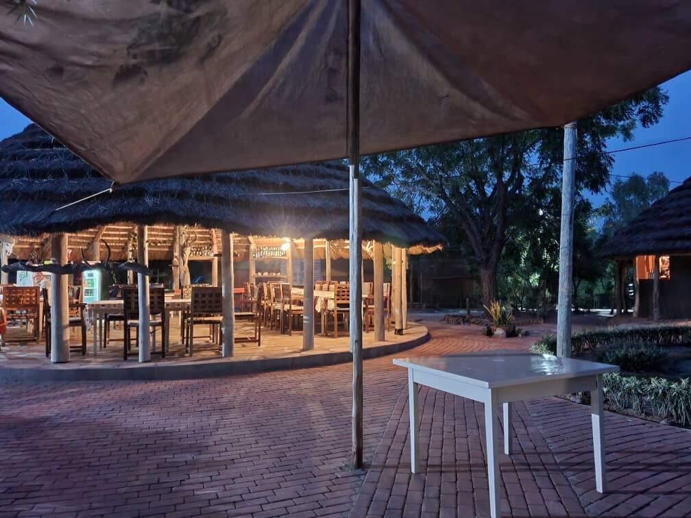 An outdoor dining area with tables and chairs under a large umbrella, surrounded by brick flooring and trees.