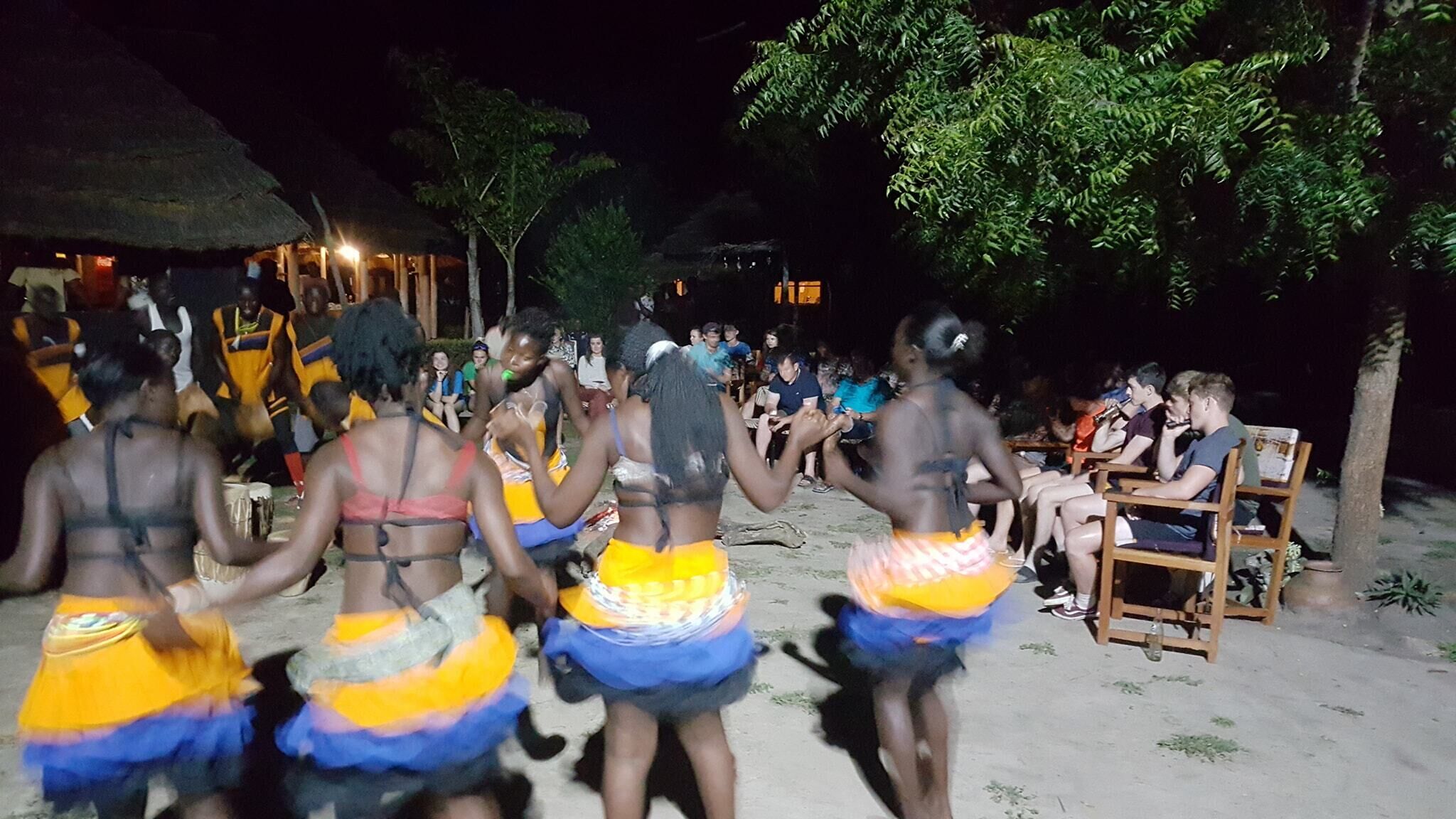 Um grupo de pessoas vestidas com trajes típicos dançando em ambiente externo, com uma construção de colmo ao fundo.