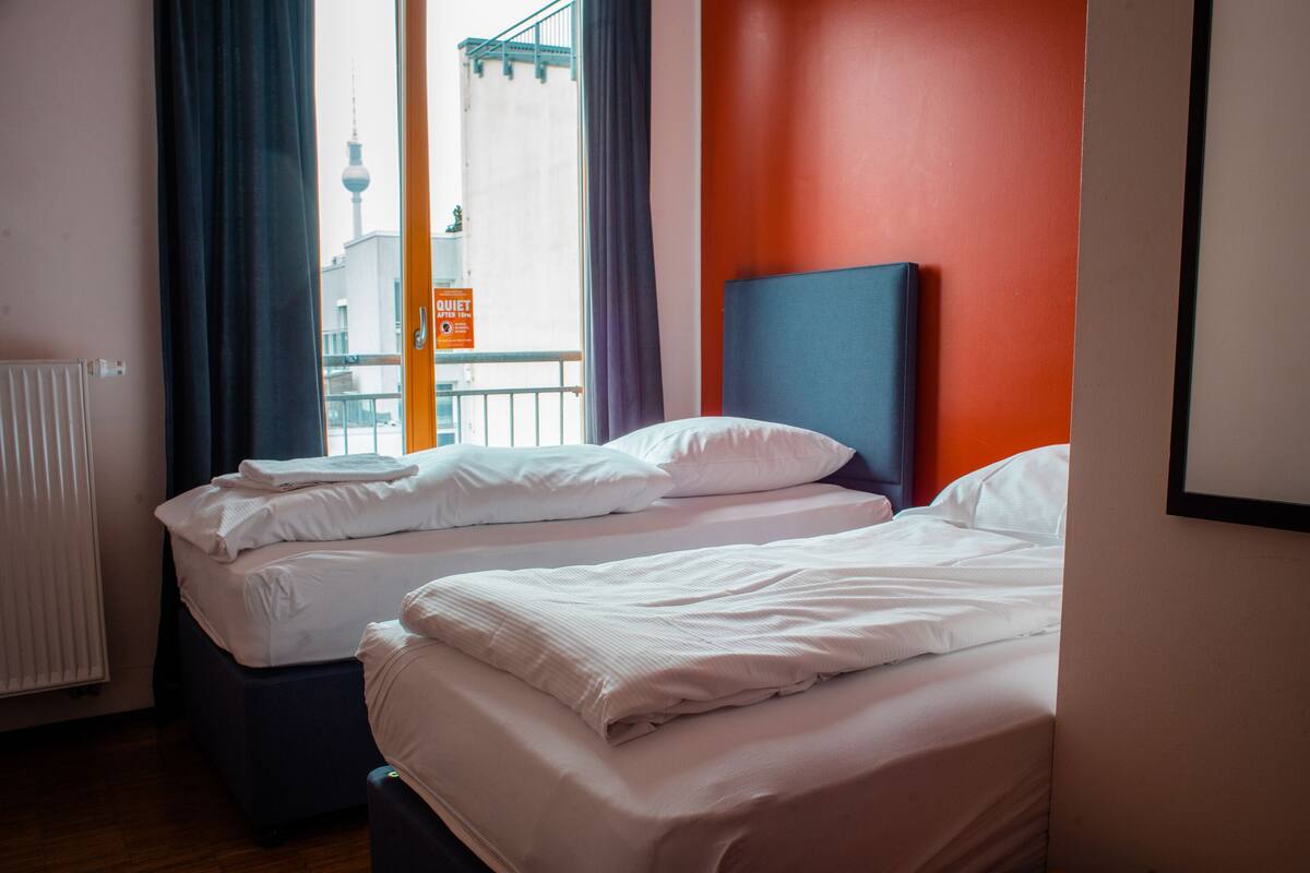 A hotel room with two beds, a view of a cityscape, and a red accent wall.