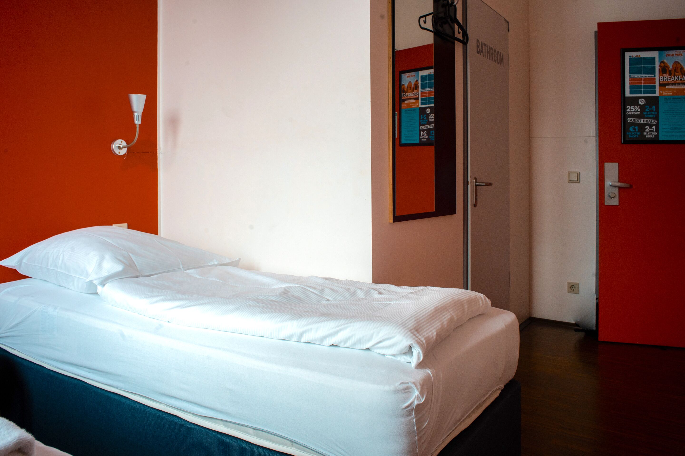 Quarto de hotel com cama, porta de banheiro e luminária fixada na parede.