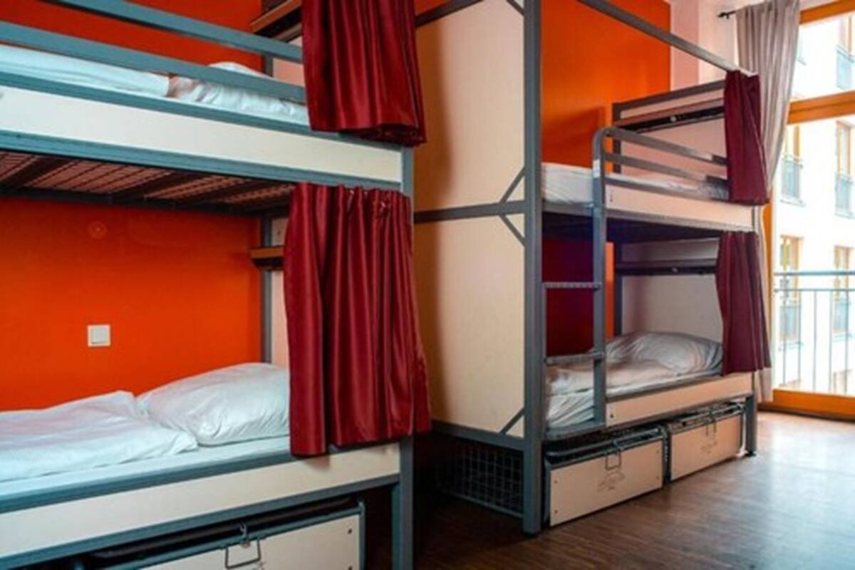 A dormitory room with bunk beds, red curtains, and a window with a view of buildings.