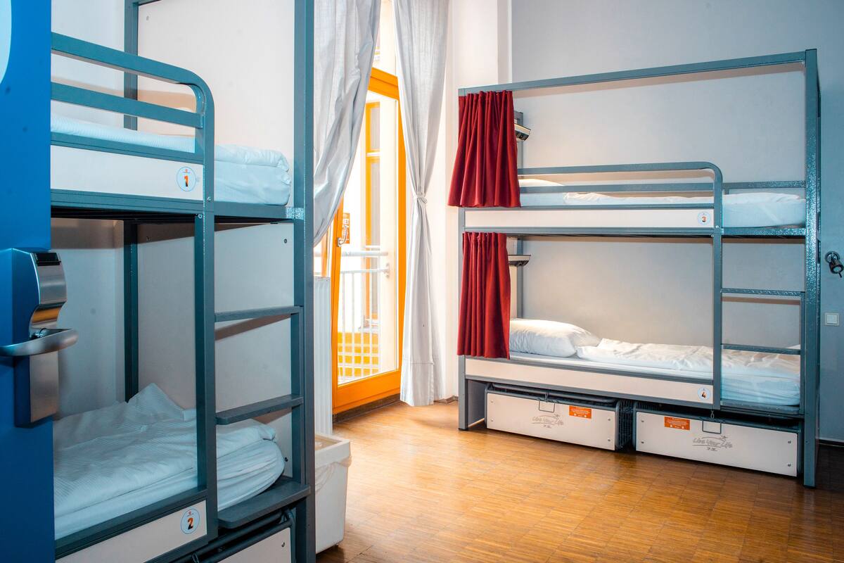A dormitory room with bunk beds, a small desk, and a window with curtains.