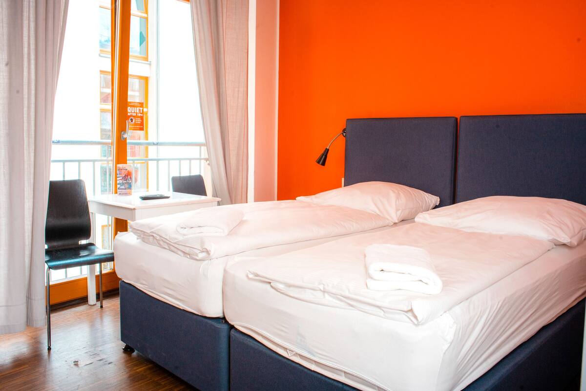 A double bed with white linens, a desk with a chair, and a window with curtains.