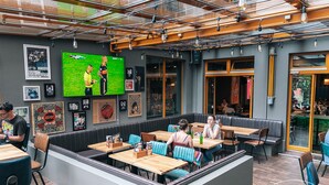 Un restaurante con cabinas y mesas, una pantalla grande mostrando un partido de fútbol y varias fotos enmarcadas en las paredes.