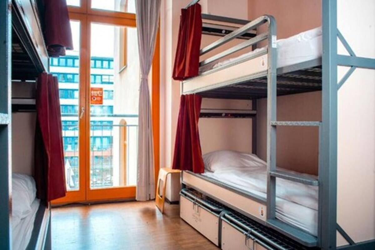 A dormitory room with bunk beds, a window with curtains, and a view of buildings outside.