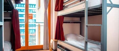 Une chambre de dortoir avec des lits superposés, une fenêtre avec des rideaux et une vue sur les bâtiments à l’extérieur.