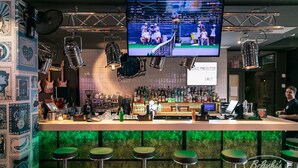 Un bar con una televisión de pantalla plana mostrando un evento deportivo, una barra con taburetes y una pared con varios carteles.