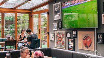 Un restaurante con un patio acristalado, mesas con sillas y una gran pantalla de televisión que muestra un partido de fútbol.