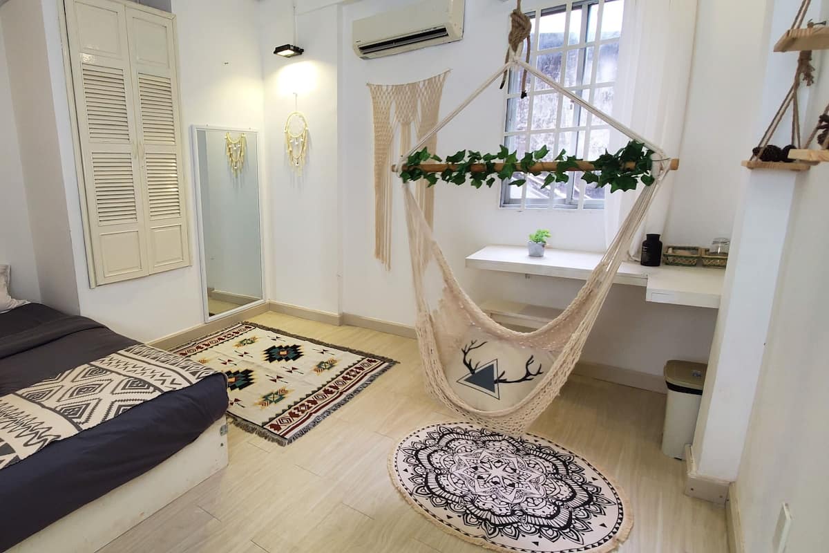 A bedroom with a bed, a hanging chair, a window, and a small table with a plant.
