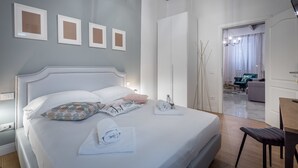 A neatly made bed with pillows and towels, a wooden floor, a white wardrobe, and a view into the living area through an open door. - Niccolò Florence Experience (Florence)