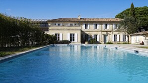 Une grande piscine rectangulaire à l’eau d’un bleu limpide, entourée d’une pelouse bien entretenue et d’un bâtiment doté d’un toit en tuiles et de volets blancs.