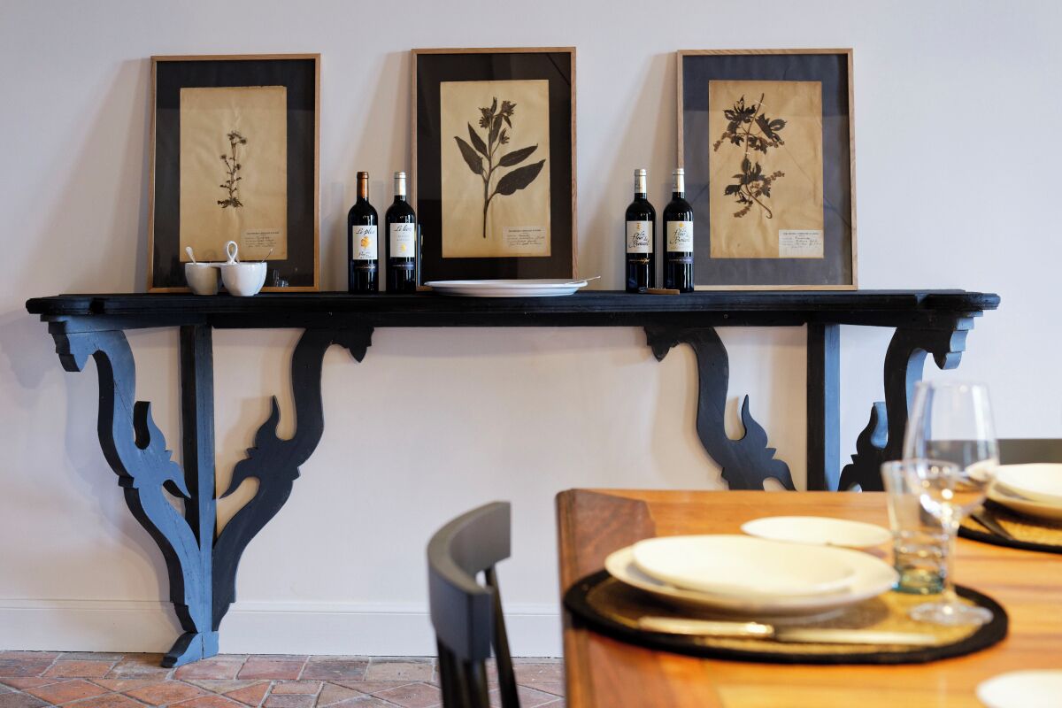 A dining area with a black ornate table, framed botanical prints on the wall, and bottles of wine.