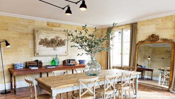 Un coin repas avec une table en bois, des chaises en osier, un vase avec des branches et un grand miroir.