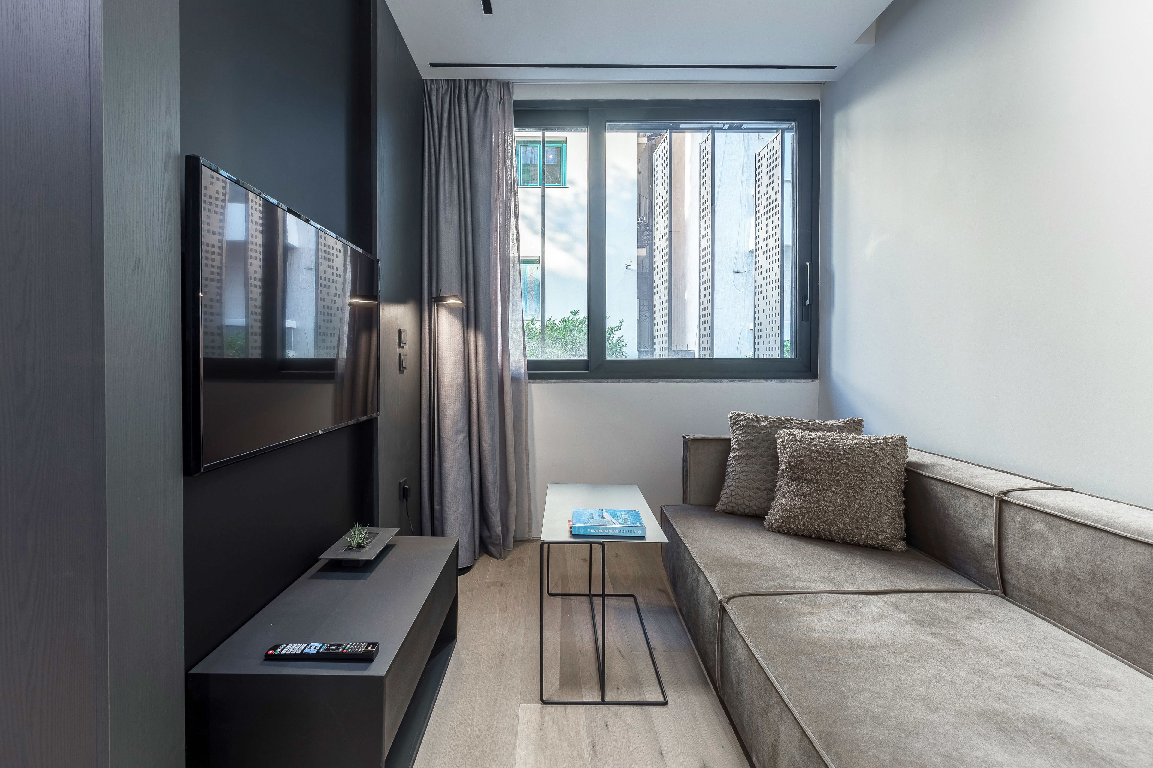 A modern living room with a sofa, a TV mounted on the wall, a small table, and a window with a view of buildings.