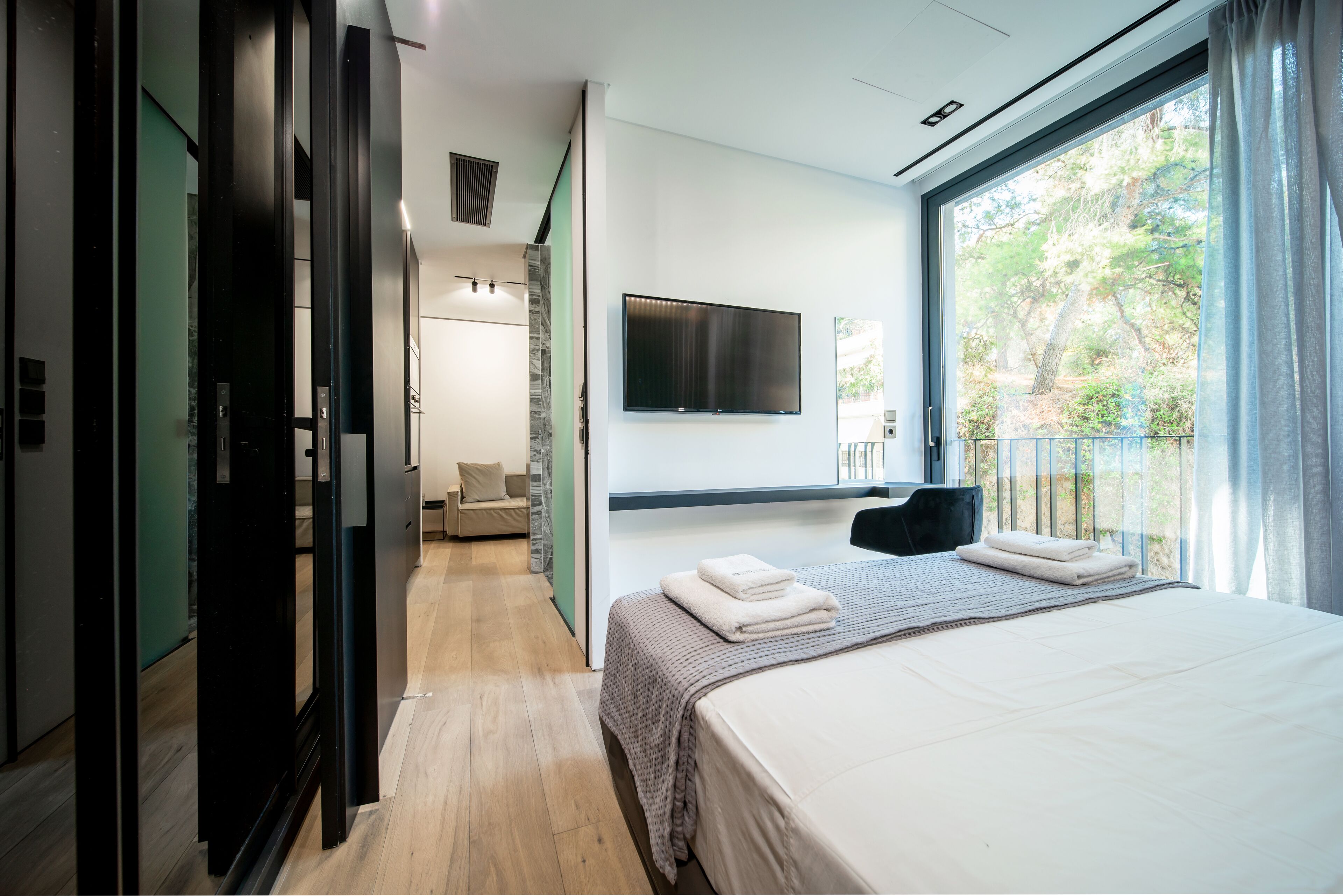 A modern hotel room with a large bed, a flat-screen TV mounted on the wall, and a balcony with a view of trees.