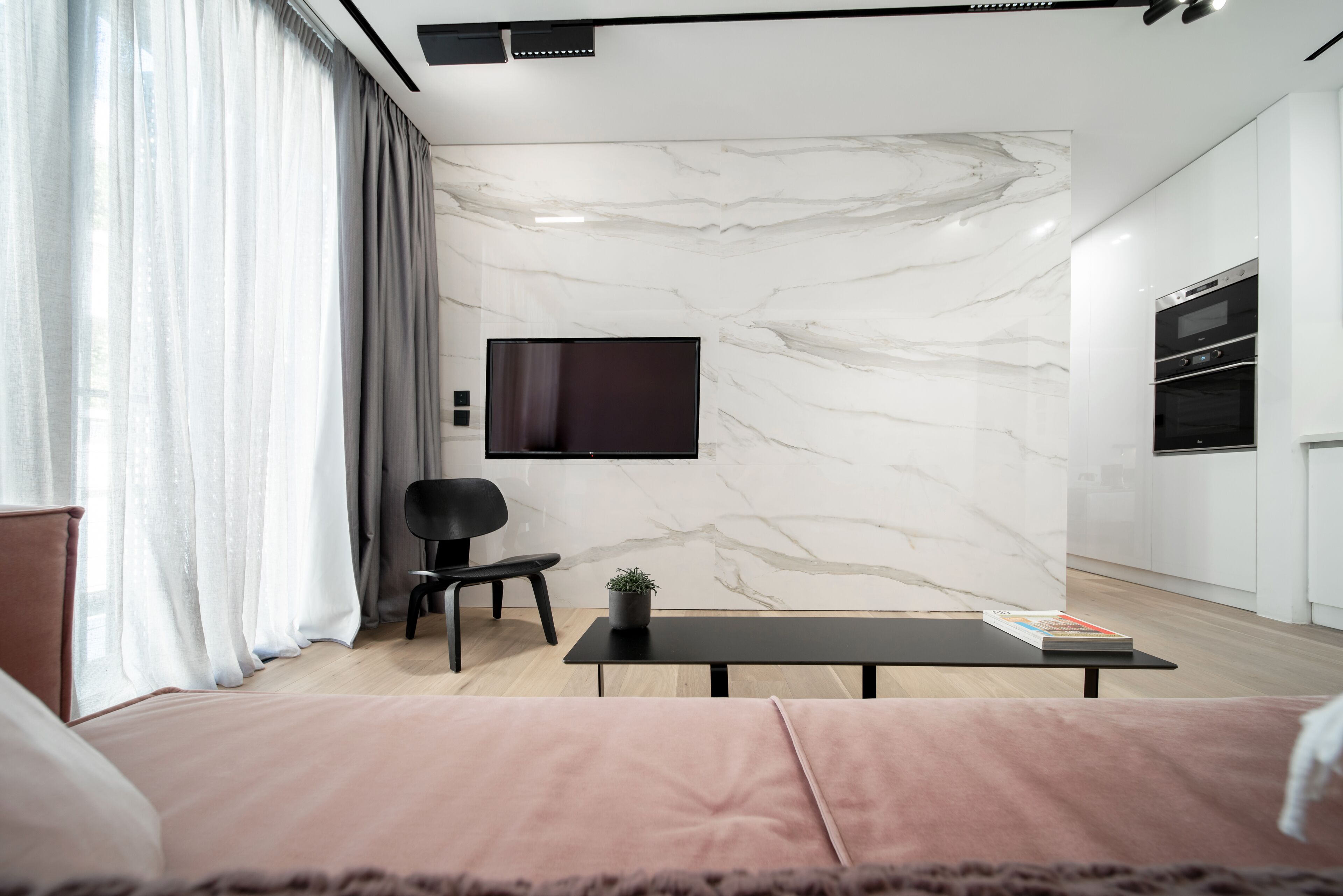 Una sala moderna con un televisor de pantalla plana montado en la pared, un sillón negro, una mesita con una planta y un sofá con funda rosa.