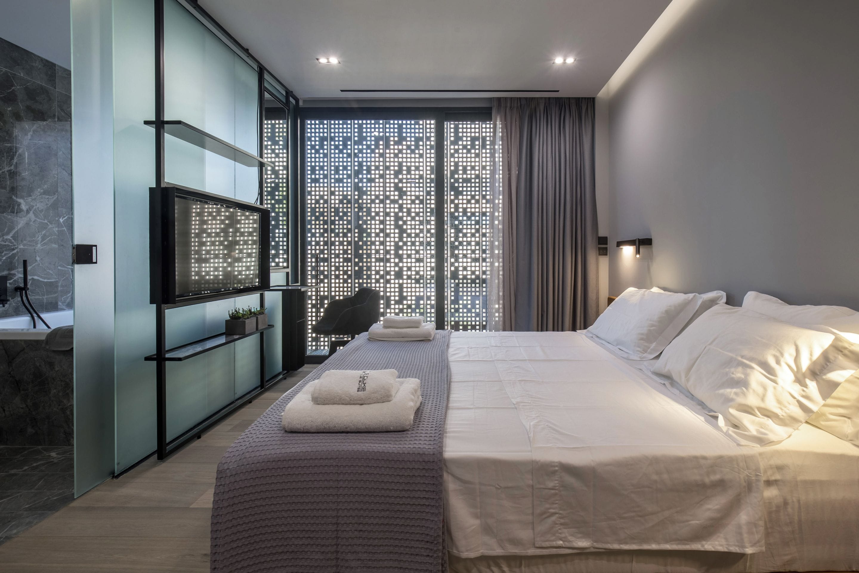A modern bedroom with a large bed, a TV, a desk, and a view through a patterned window.