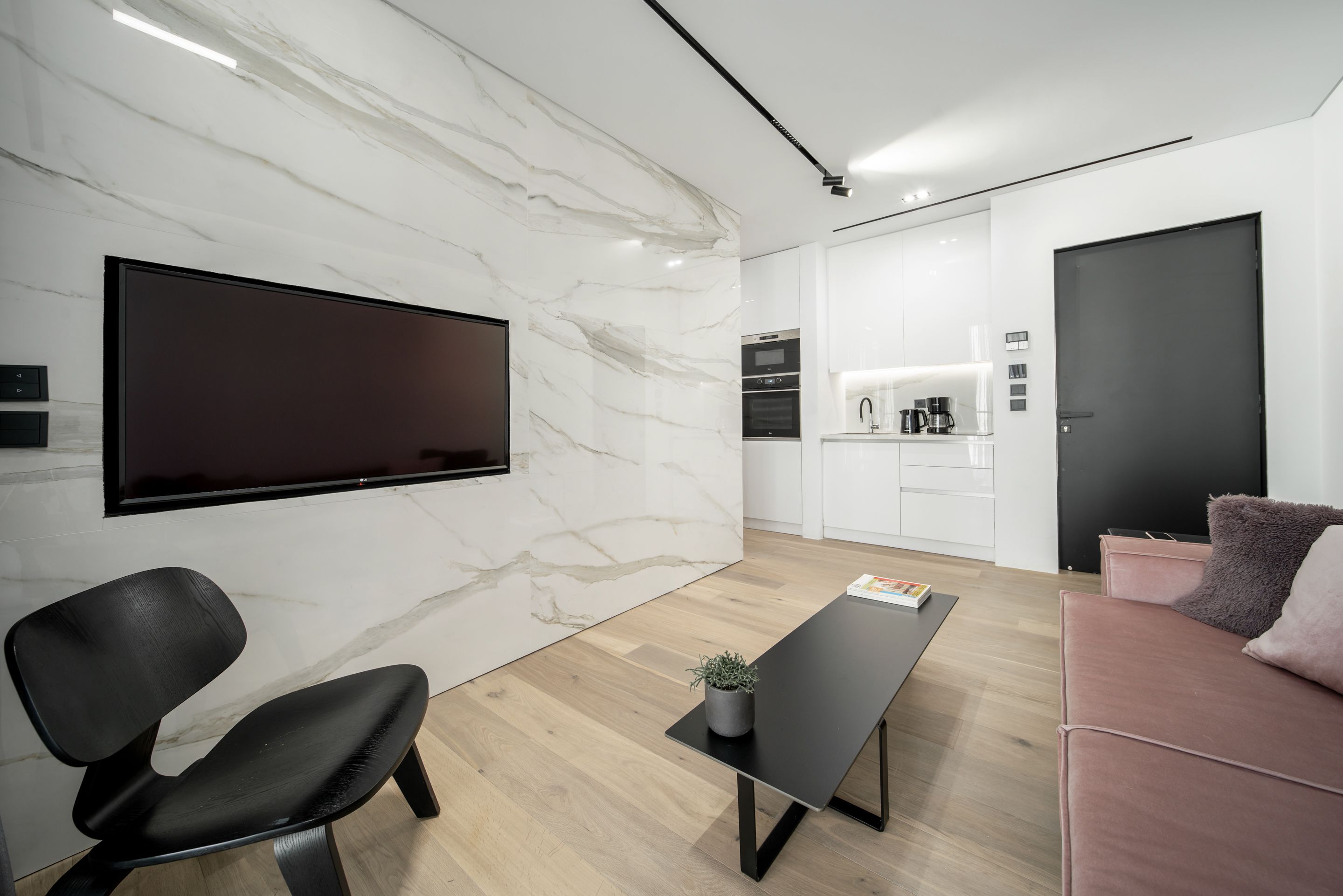 Un salón moderno con un televisor de pantalla plana, una butaca negra, una mesa de centro con una planta y un sofá rosa.