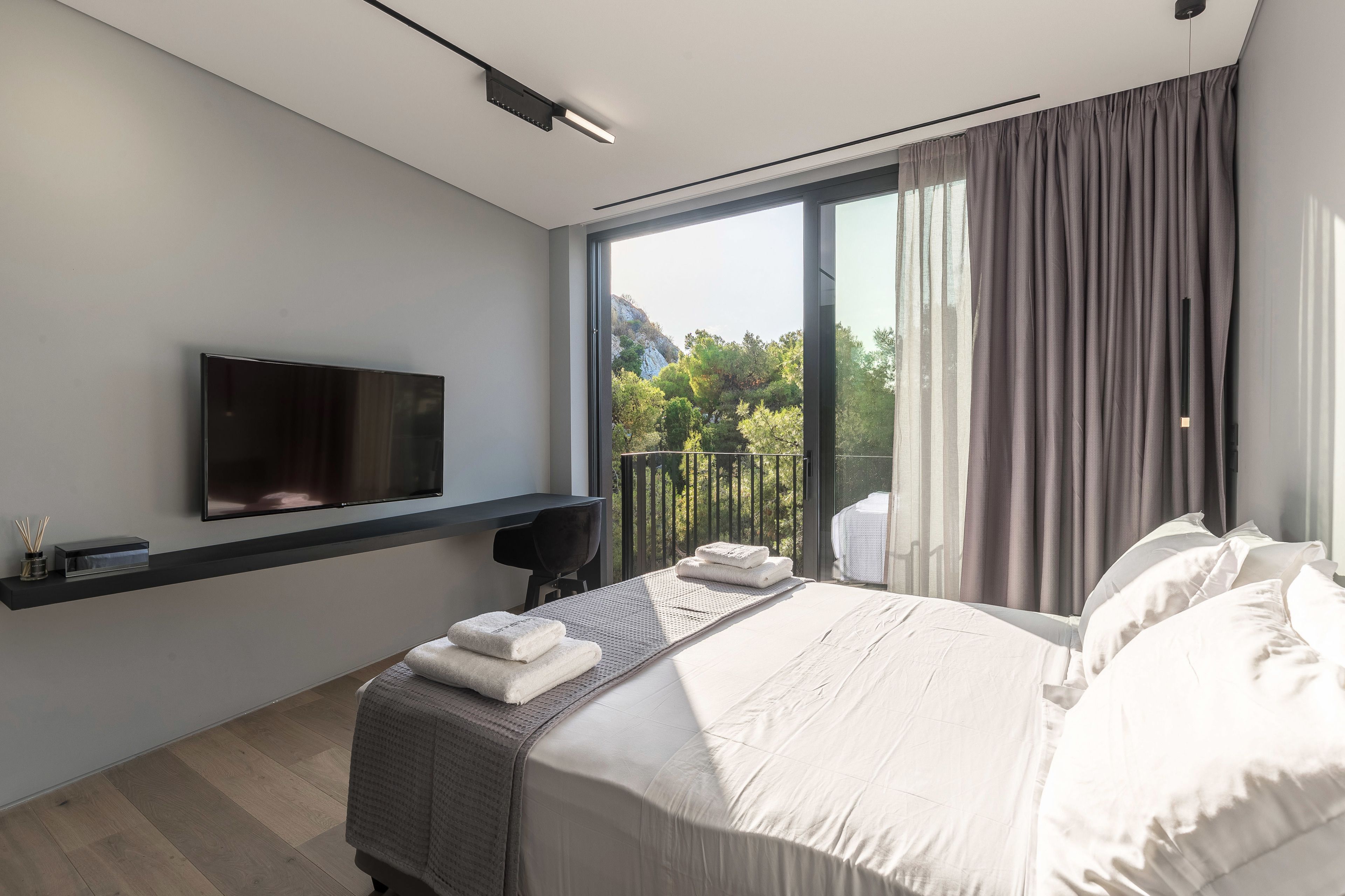 A modern hotel room with a large bed, a flat-screen TV mounted on the wall, a small desk, and a balcony with a view of trees and mountains.
