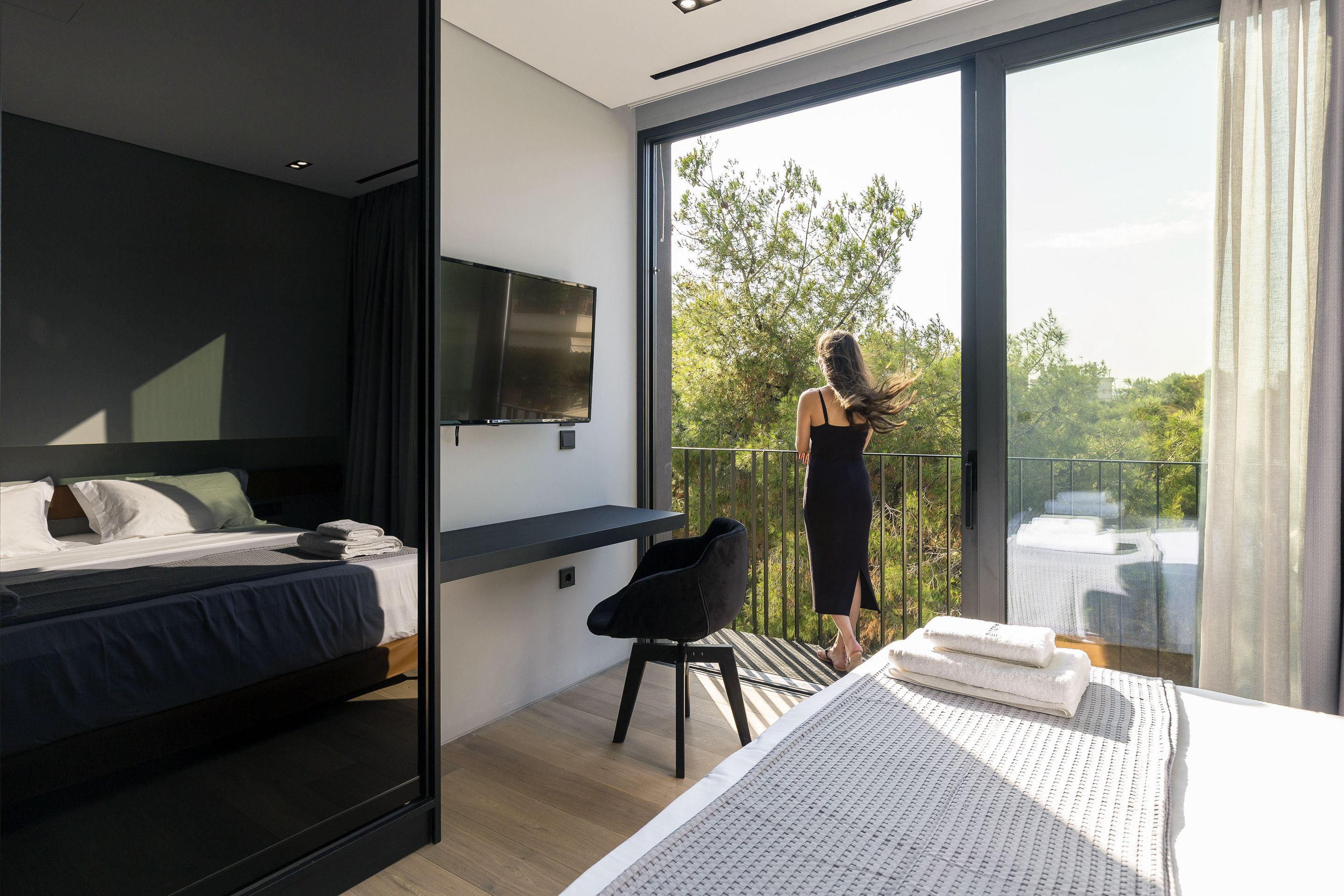 A modern bedroom with a bed, a TV, and a balcony with a view of trees.