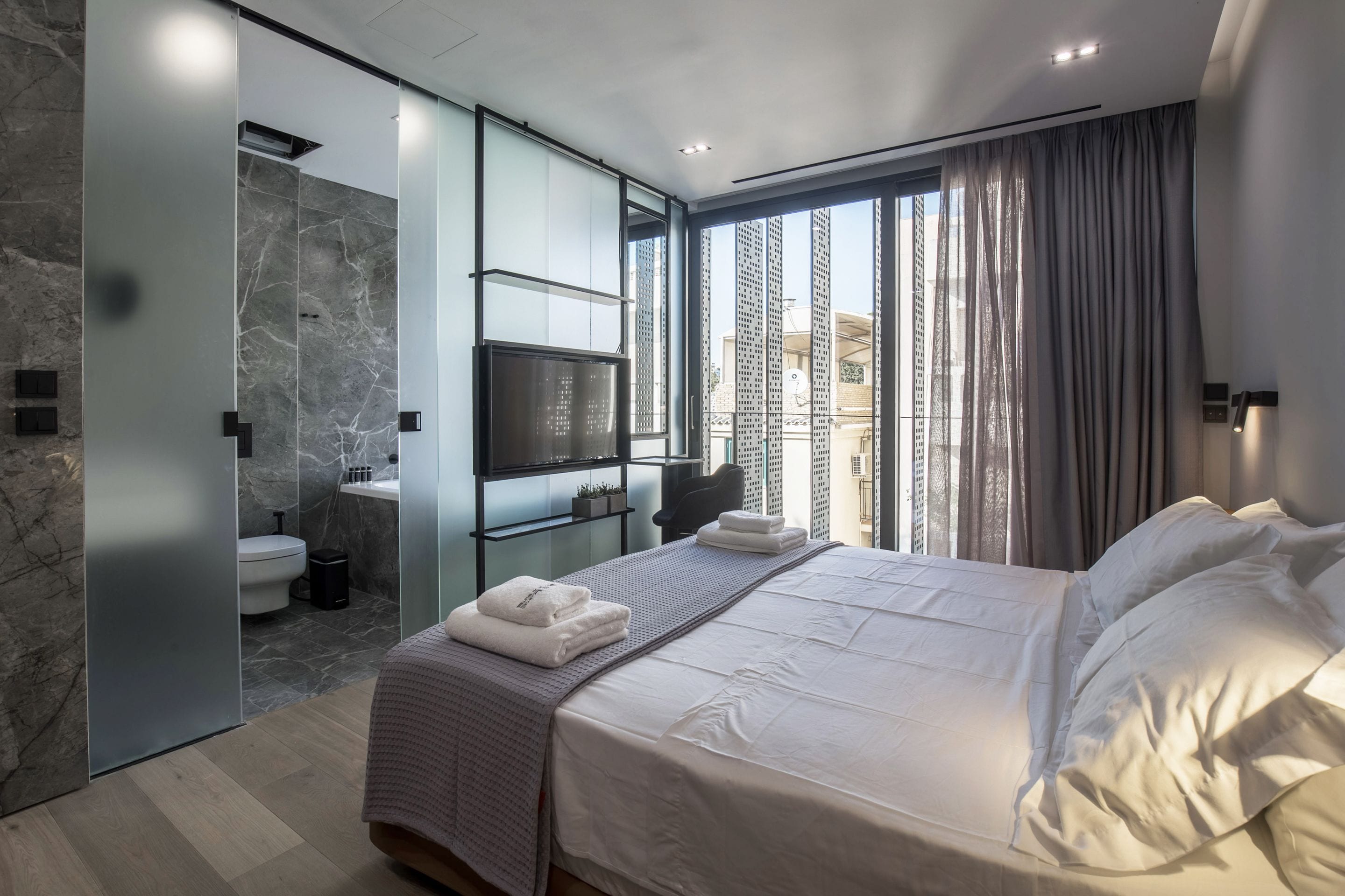 Ein modernes Schlafzimmer mit einem großen Bett, einem Fernseher, einem Badezimmer mit Toilette und Waschbecken sowie einem Blick über die Stadt durch das Fenster.