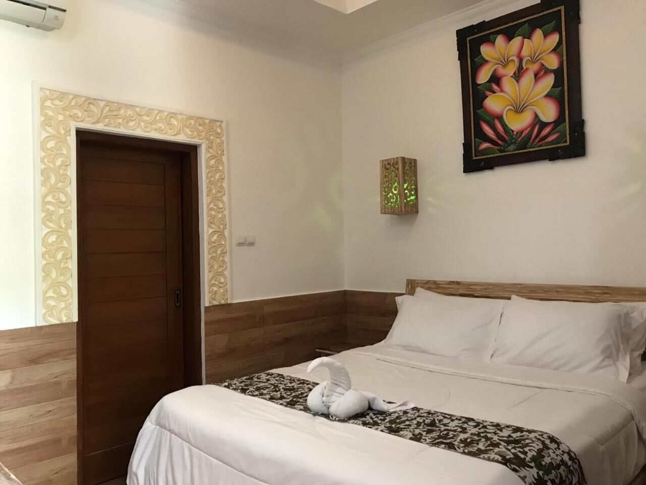 Une chambre d’hôtel avec une porte en bois, un lit avec une literie blanche, un panneau décoratif mural et une œuvre d’art florale encadrée.