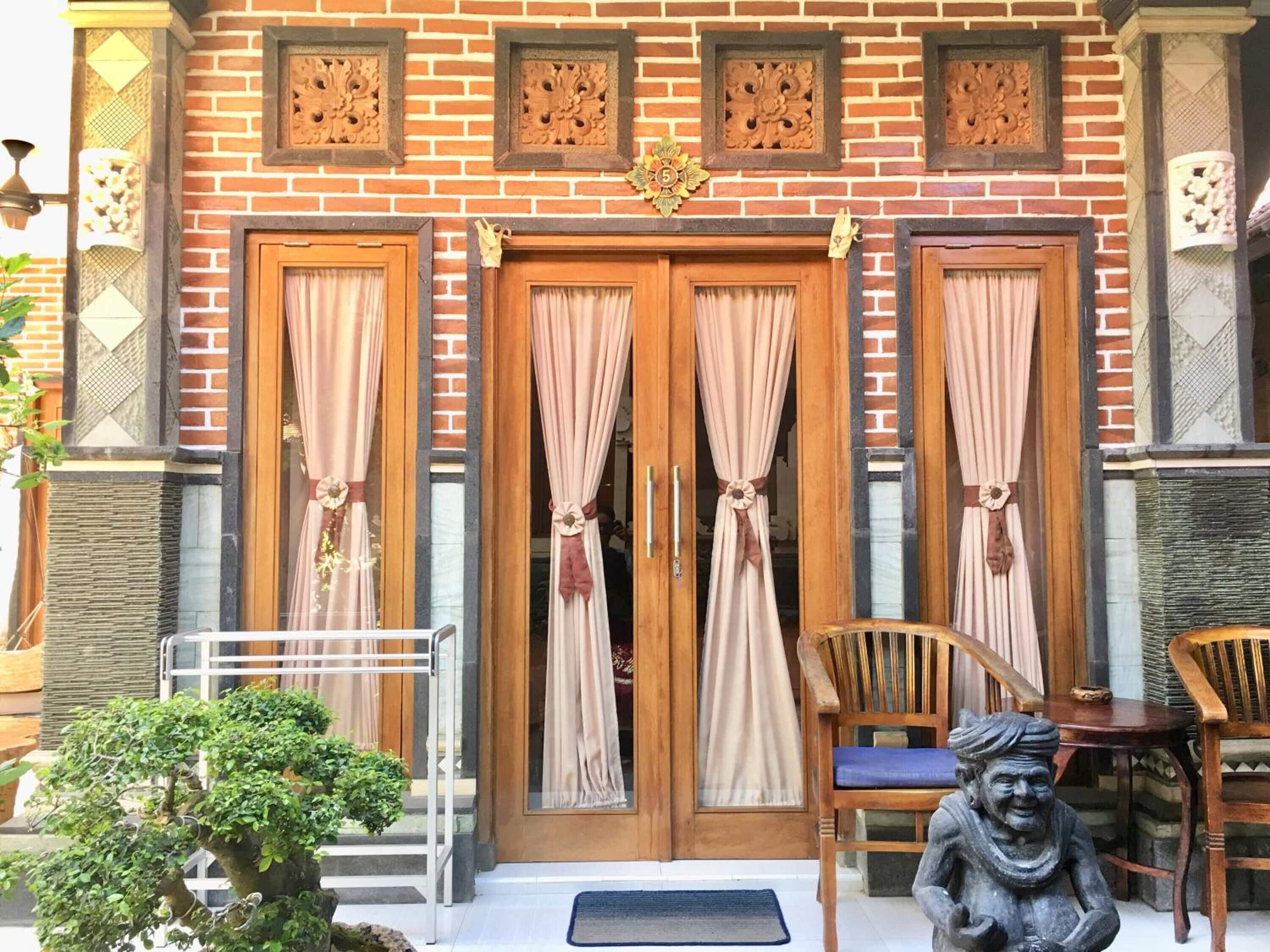 A brick building with three wooden doors, each with curtains. There is a small statue and a chair outside.
