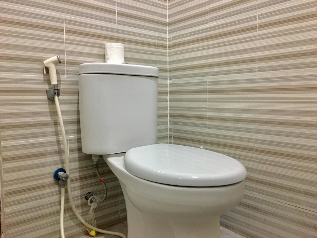 Un inodoro con tanque y tapa blancos, conectado a una tubería y una manguera fijadas a la pared.