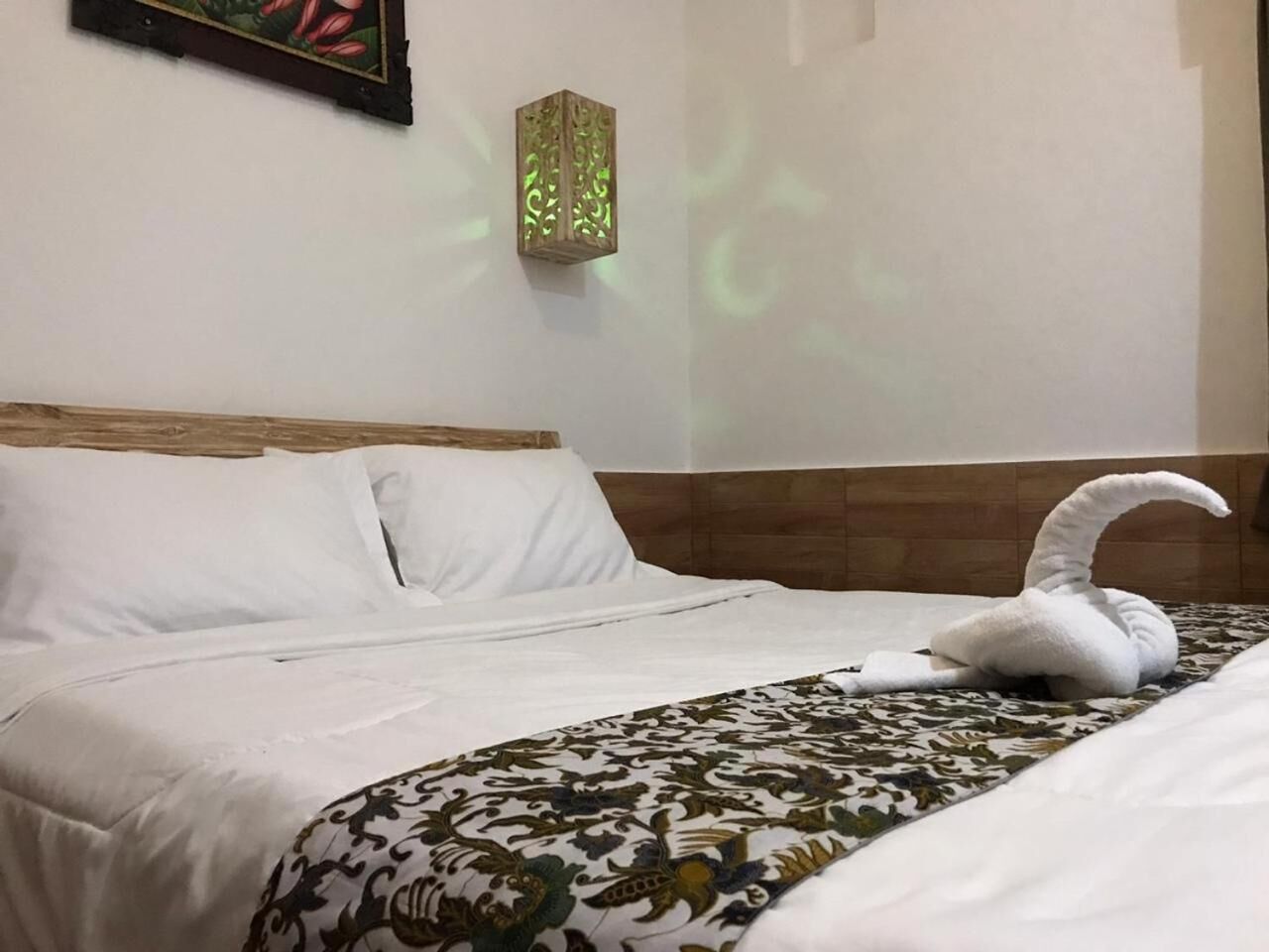 A hotel room with a neatly made bed, a decorative wall lamp, and a framed artwork above the bed.