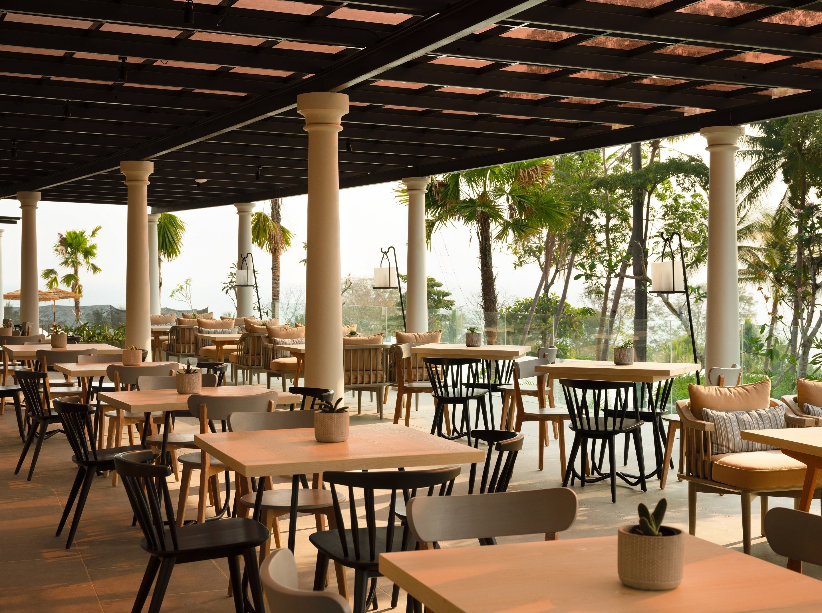 An outdoor cafe with multiple tables and chairs, large white columns, and a view of palm trees and mountains.