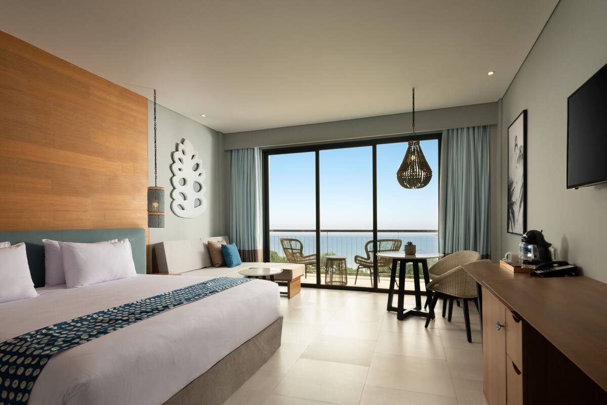 Habitación de hotel moderna con una cama grande, un balcón con vistas, un rincón de comedor y un televisor.