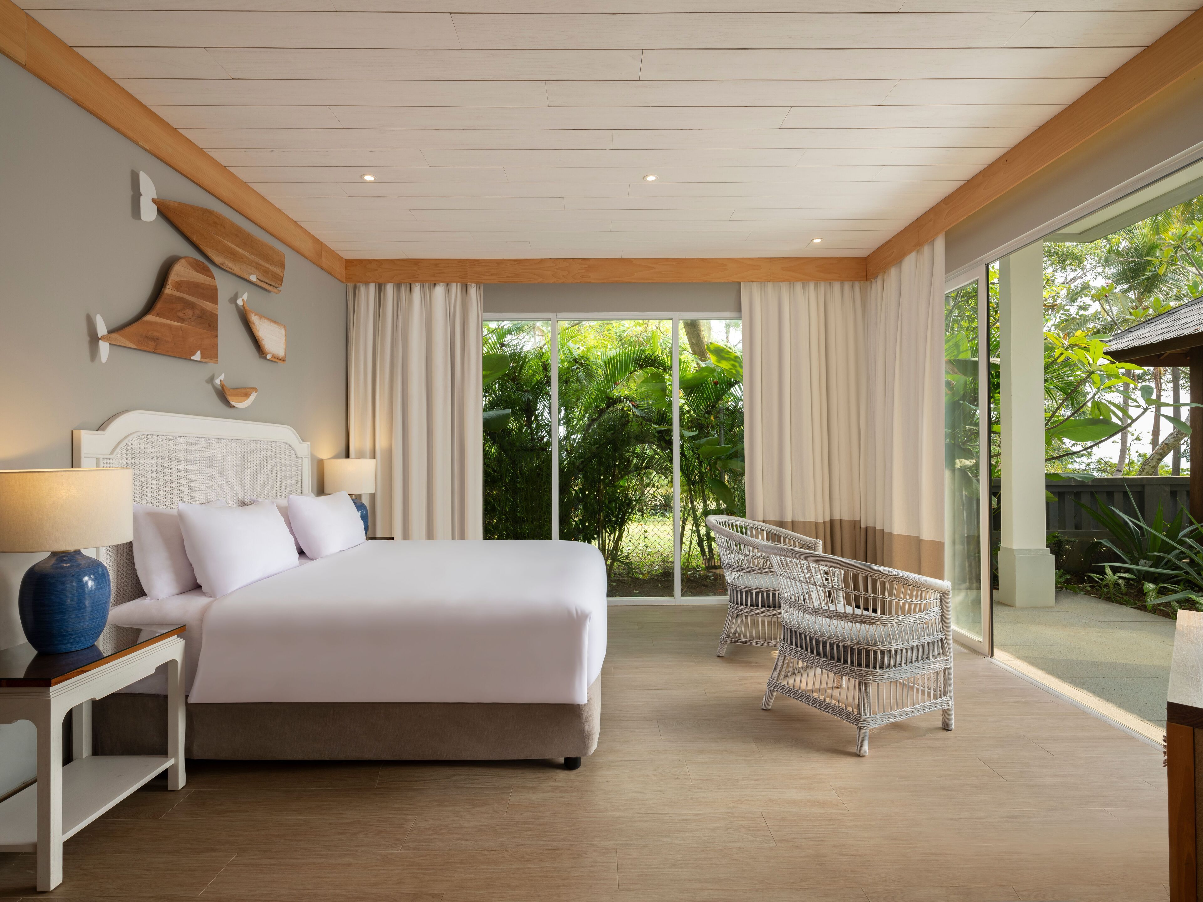 A bedroom with a large bed, bedside tables, a wicker chair, and a view of greenery through the window.
